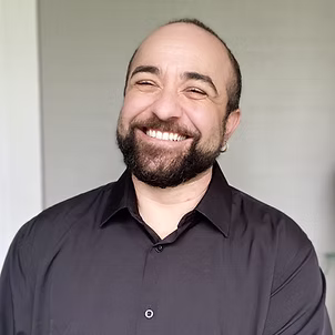 Headshot of Dylan smiling and laughing. They have short hair, a beard, and a black button-down shirt.