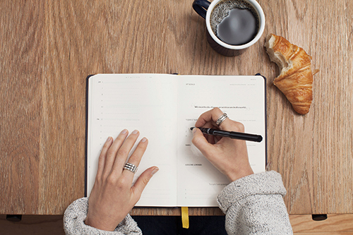 Overhead shot of hands on a planner on a wood desk, with a pen in the right hand and coffee and a croissant nearby