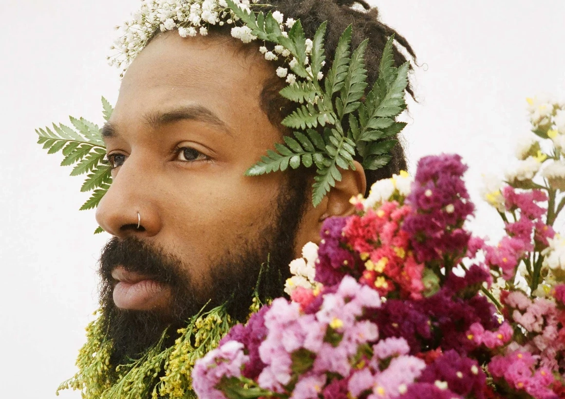 A bearded Black man, adorned with leaves and flowers, gazes off into the distance