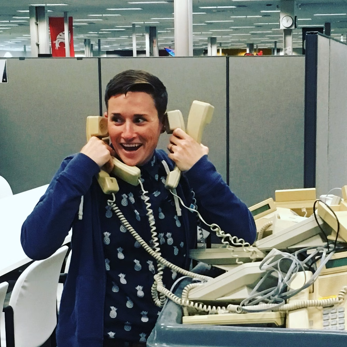Kat stands holding 5 phones to their ears in a silly way of depicting busyness. The phones are old school corded landlines.