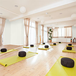 Bright yoga studio with white walls, wooden floor, yellow mats, black cushions, and large windows with curtains.