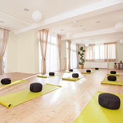 Bright yoga studio with off white walls, yellow mats and black round cushions arranged on wooden floor.