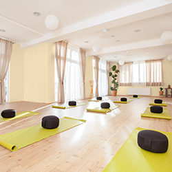 Bright yoga studio with yellow walls, wooden floor, green mats, and black meditation cushions near large windows.