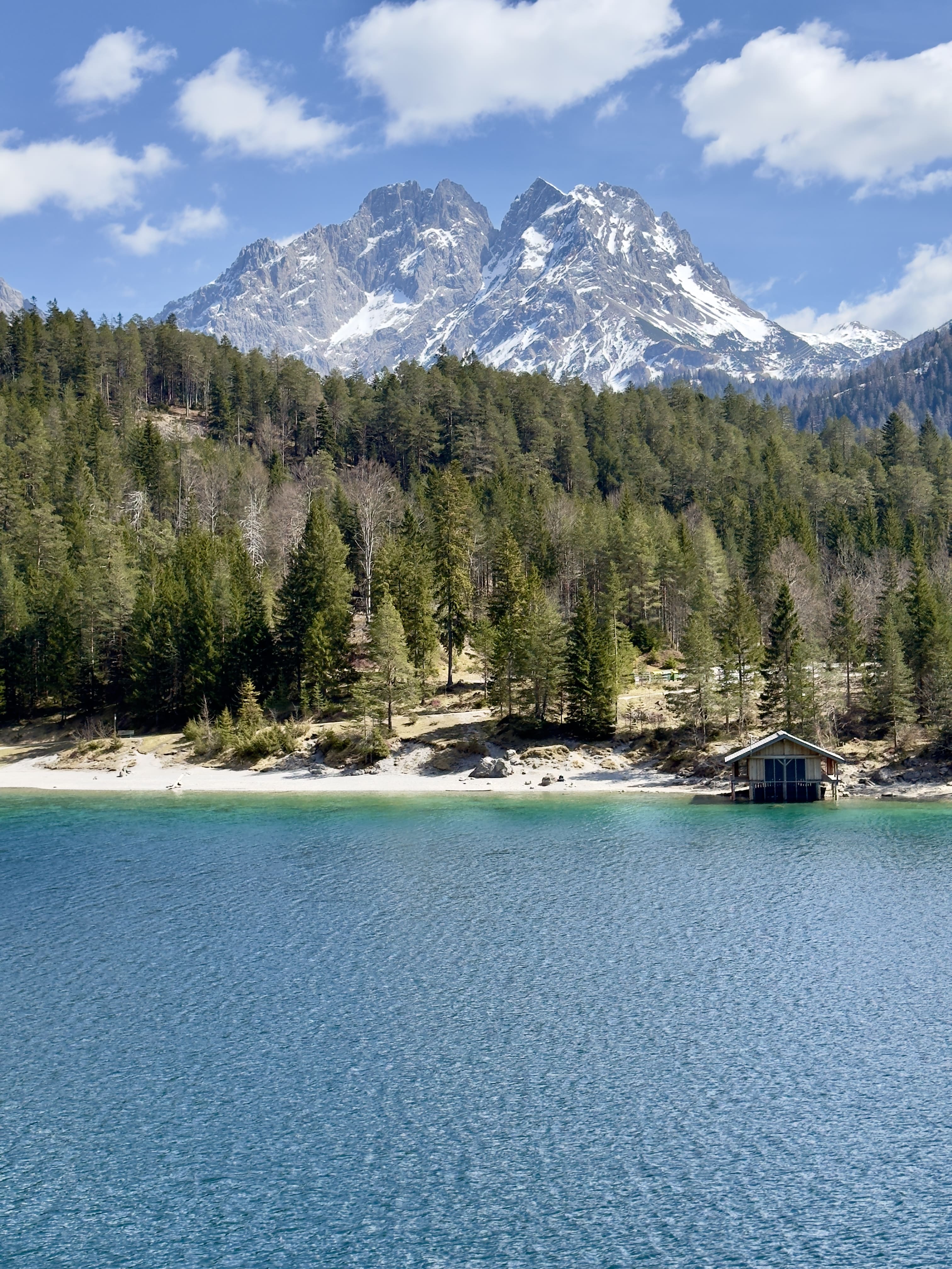 Clear blue lake with a small wooden boathouse on shore, dense forest, and snow-capped mountains under a partly cloudy sky.