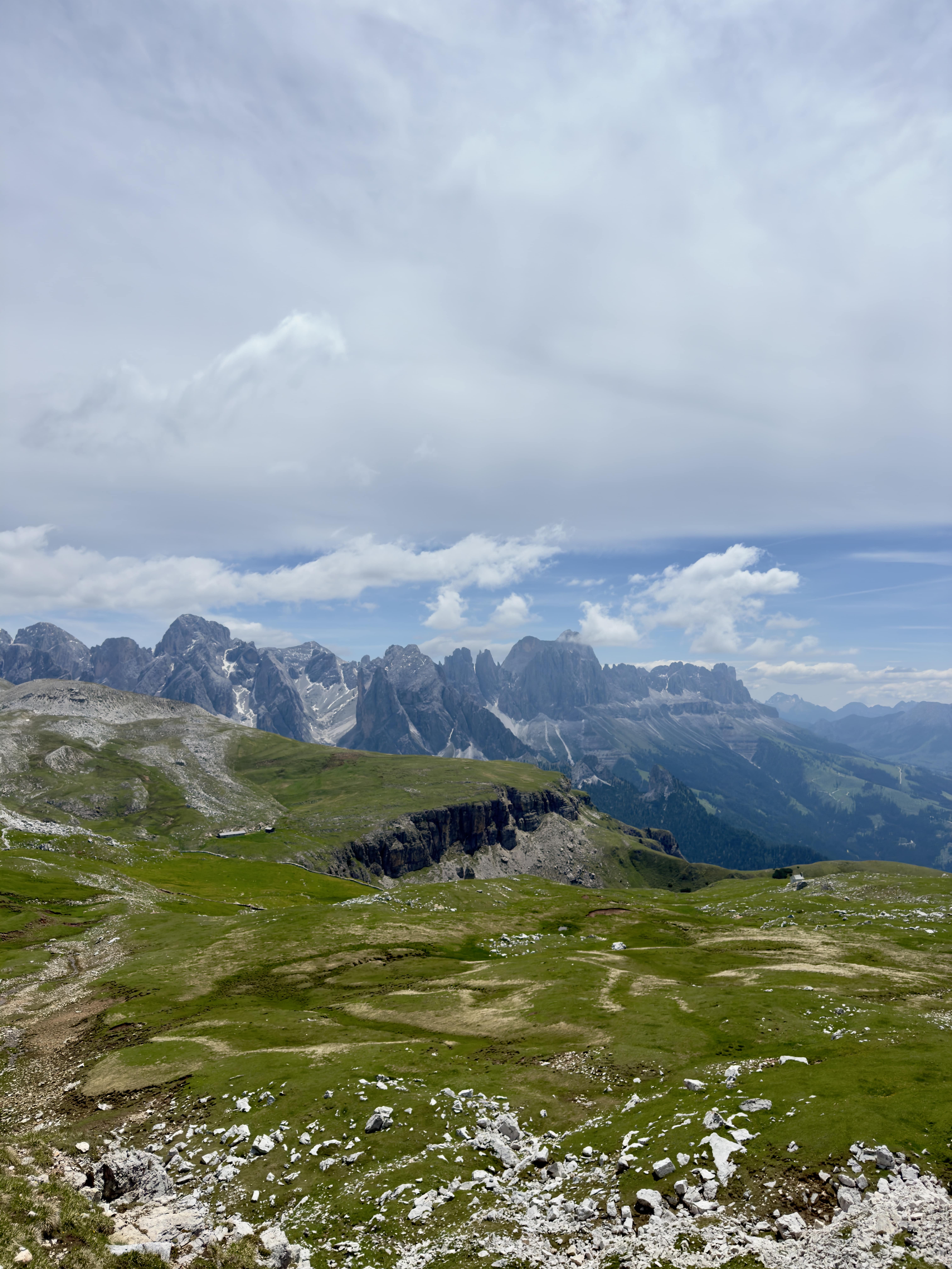 Vast green meadow with rocky patches in front of a mountain range under a partly cloudy sky.
