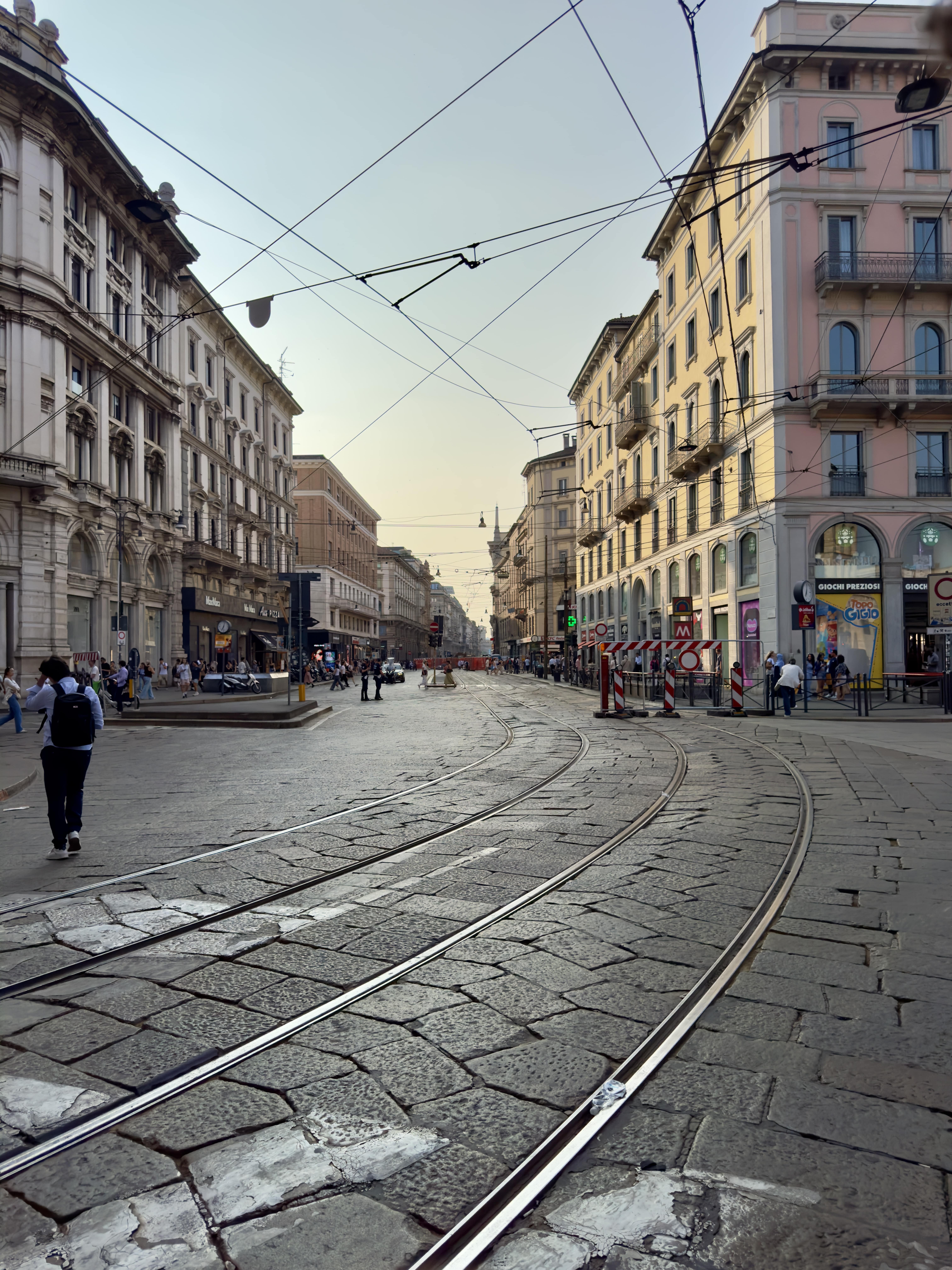 Urban street with tram tracks curving between historic buildings and pedestrians on a clear day.