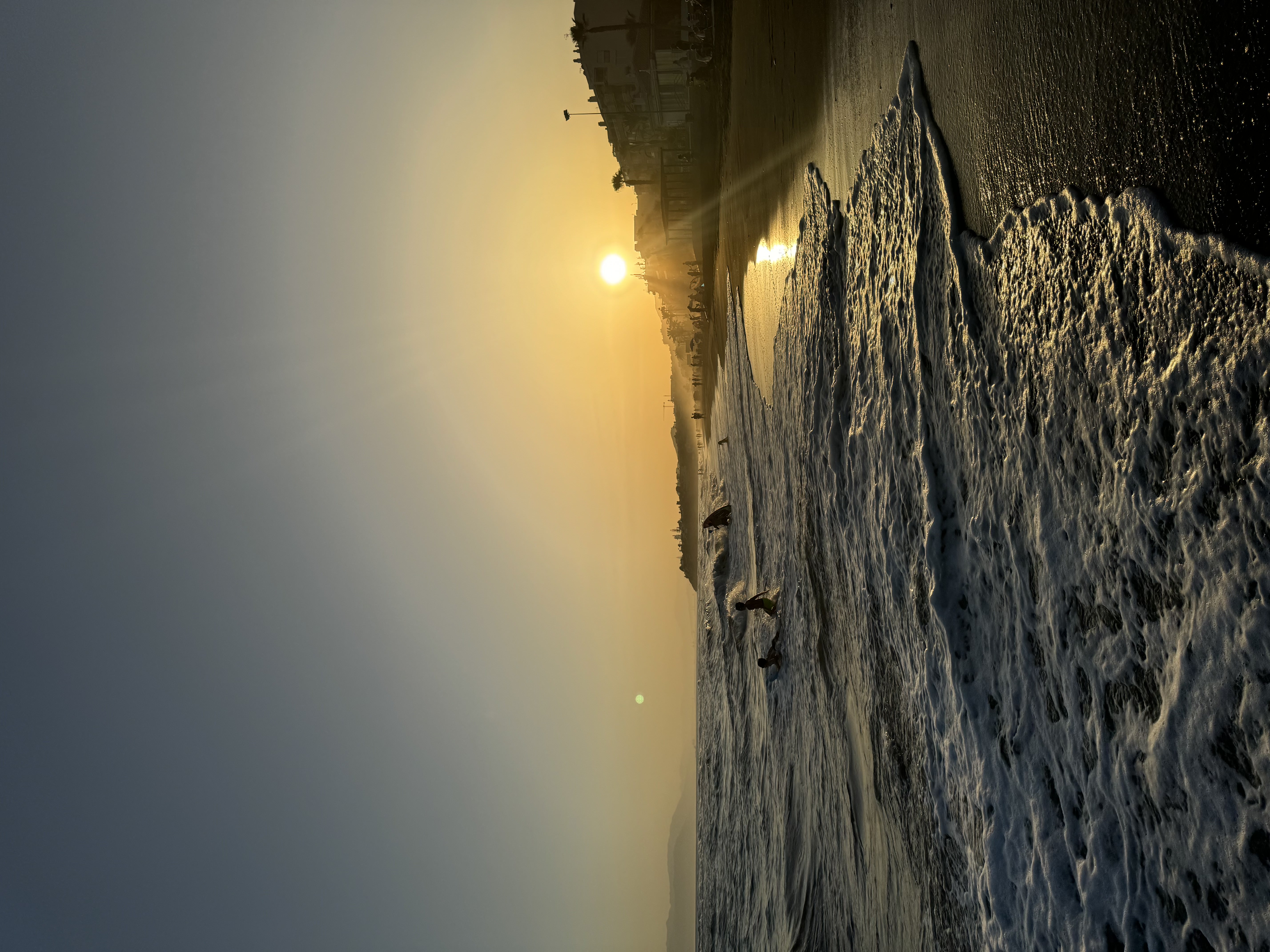 Sunset over a beach with waves washing onto the shore and people playing in the water.