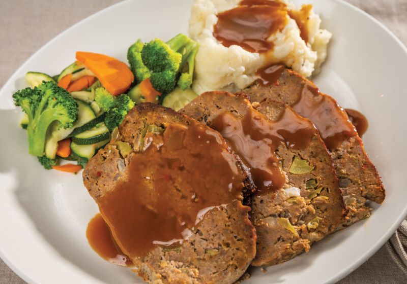 A LARGE WHITE PLATE HAS A 4 BIG SLICES OF MEATLOAF WITH MIXED VEGGIES AND MASHED POTATOES, ALL SMOTHERED INA BROWN GRAVY.