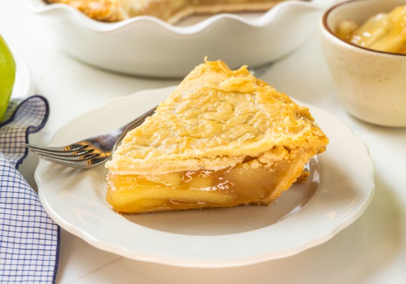 A SMALL WHITE PLATE HAS A FORK AND  A PEICE OF APPLE PIE ON IT.  BEHIND IT IS A BLURRY IMAGE OF THE WHOLE PIE THE PIECE CAME FROM.