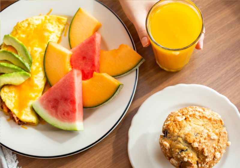 A BLACK RIMMED PLATE HAS A OMELETE  WITH WATERMELON AND A SIDE OF CATALOUPE. ON THE OMELETE THERE IS CHEESE AND AVOCADO. NEXT TO THE PLATE THERE IOS A  PERSON HOLDING A GLASS OF ORANGE JUICE AND A THERE IS A SMALL PLATE WITH A MUFFIN ON IT