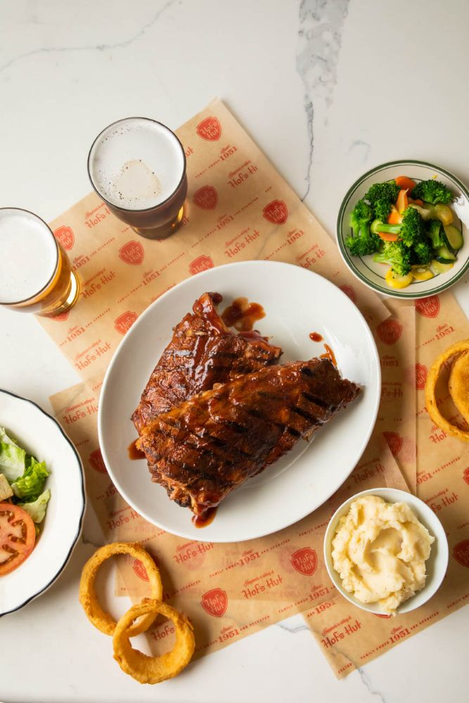 A LARGE PLATE OF RIBS WITH A SIDE OF VEGGIES, GARLIC MASHED POTATOES AND A SIDE SALAD.  AROUND THE RIBS THERE ARE A FEW ONION RINGS AND TWO PINTS OF BEER.
