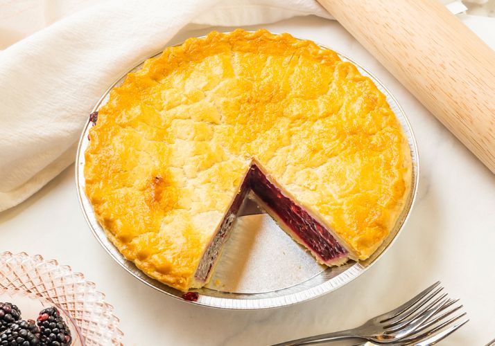 A large berry pie with a piece cut out of it. Fresh Berries on the side along with 2 silver forks and a wooden rolling pin. 