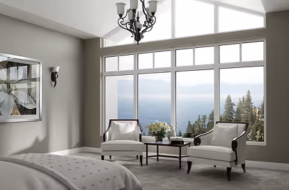 Bright bedroom with two white armchairs, a small table with flowers, large windows showing a view of trees and mountains.