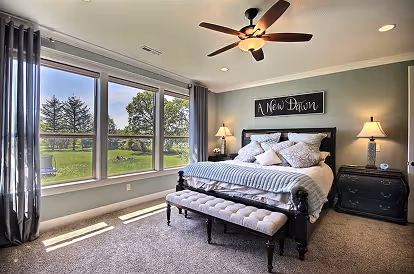 Cozy bedroom with large window, ceiling fan, bed with multiple pillows, two lamps, and bench at foot of bed.