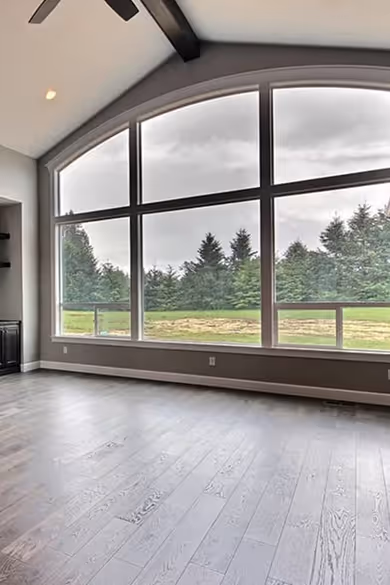 Empty room with large arched windows overlooking green trees and grass under a cloudy sky.