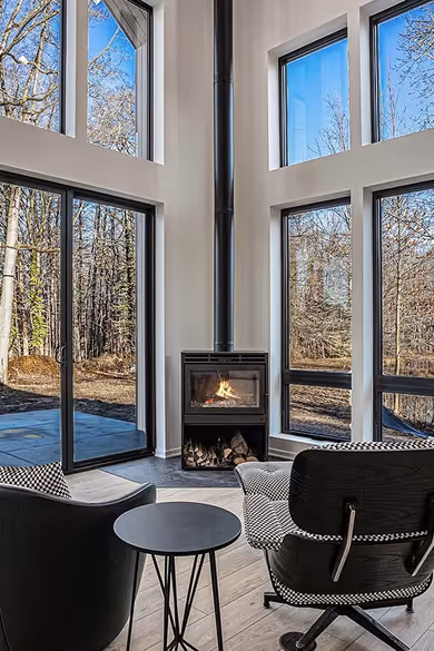 Modern living room with large floor-to-ceiling windows, a black wood stove with fire, two chairs, and a small round table.