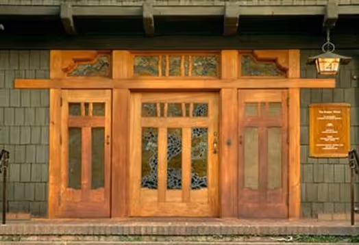 Wooden front door with decorative glass panels and side windows on a green shingle wall, with a hanging lantern and signboard on the right.