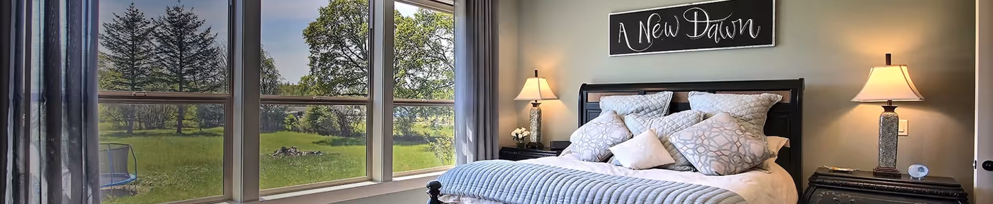 Cozy bedroom with a large bed, multiple pillows, two bedside lamps, and a window showing a green outdoor landscape.