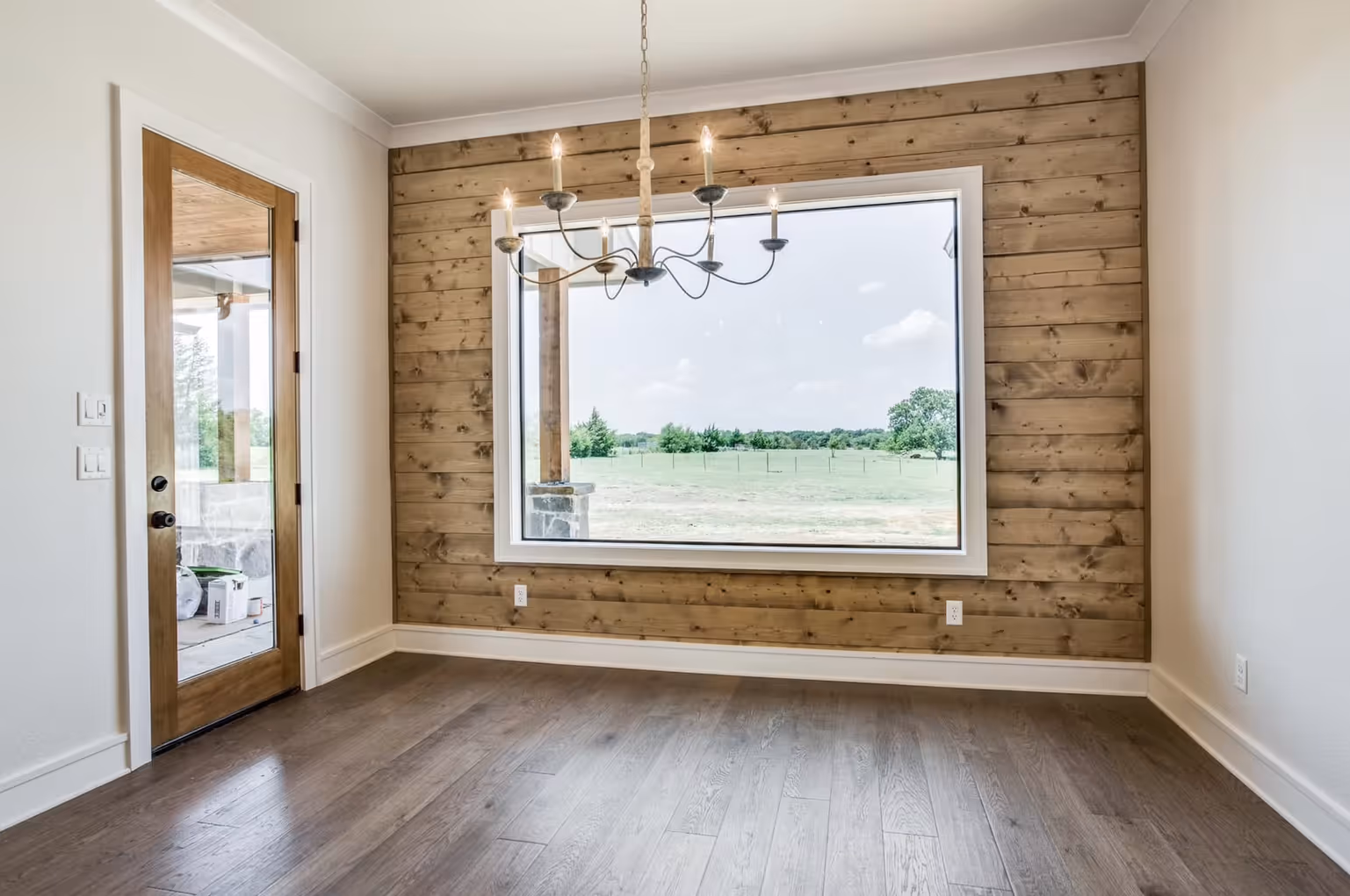 Empty room with wooden floor, wood-paneled wall featuring a large window showing a green field, a wooden framed glass door, and a chandelier with six lit candles.