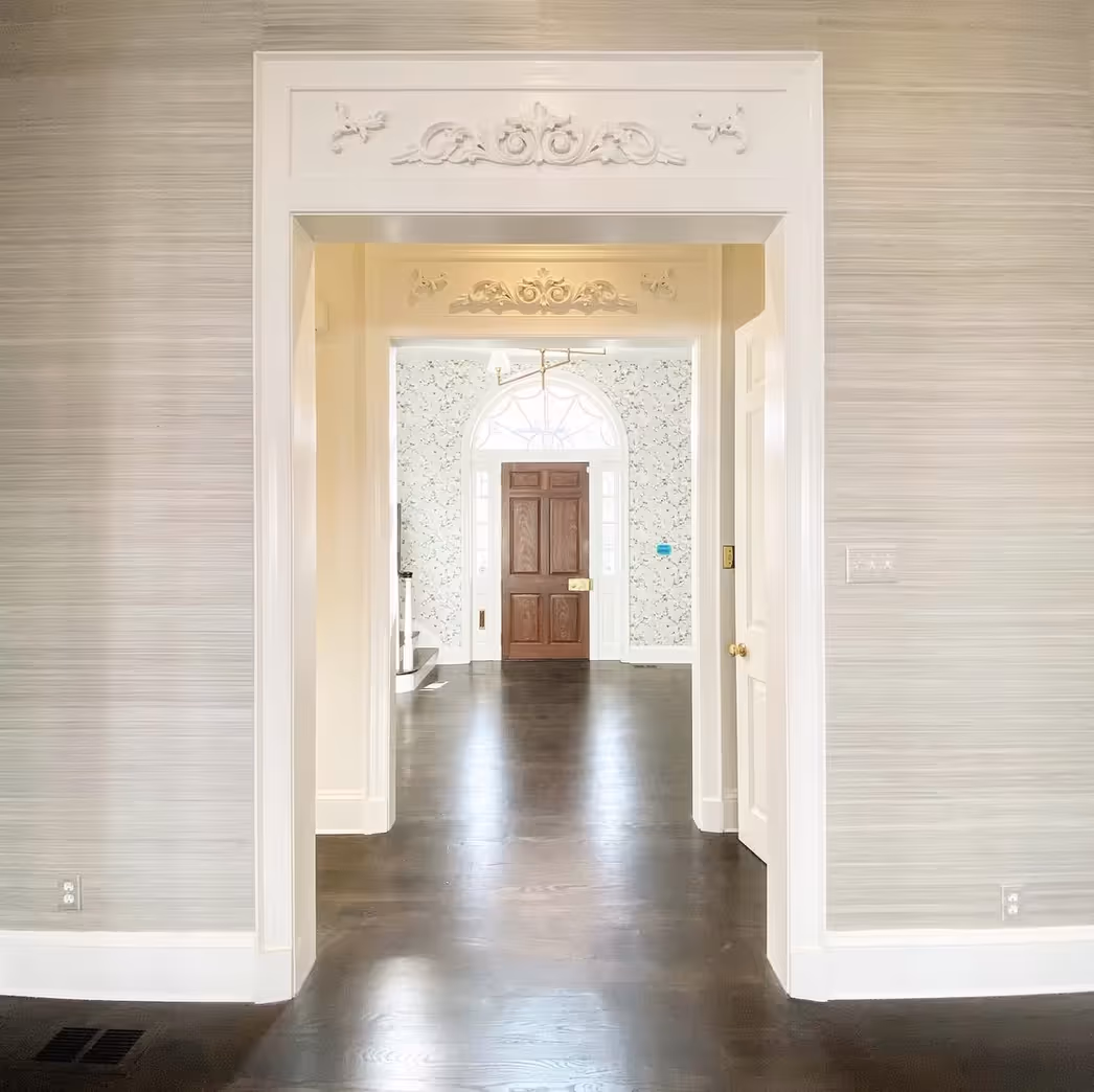 View through multiple doorways revealing a dark wooden front door with decorative white trim and floral wallpaper in the entrance hall.