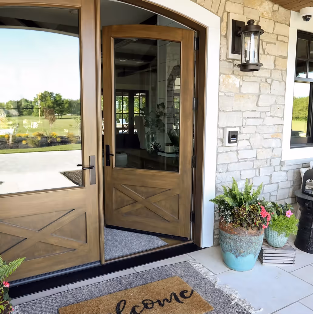 Open wooden front door with glass panels, a welcome mat, potted plants, and stone wall exterior.