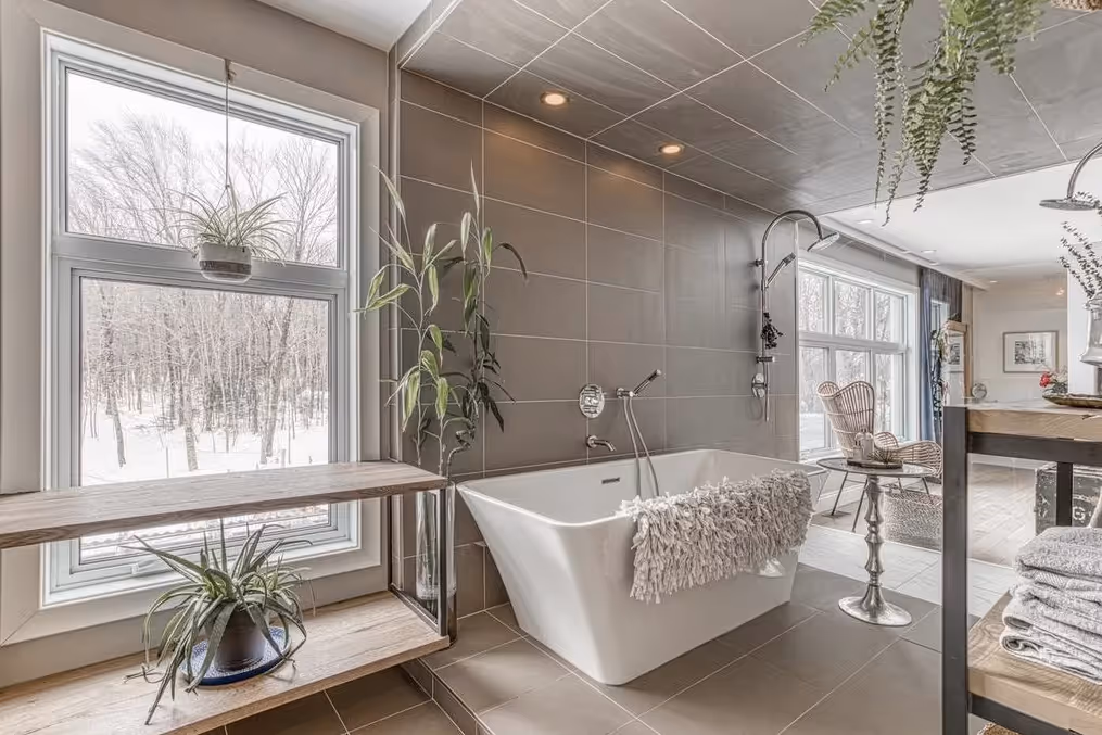 Modern bathroom with freestanding rectangular bathtub, grey tiled walls, hanging and potted plants, and large windows showing a snowy forest outside.