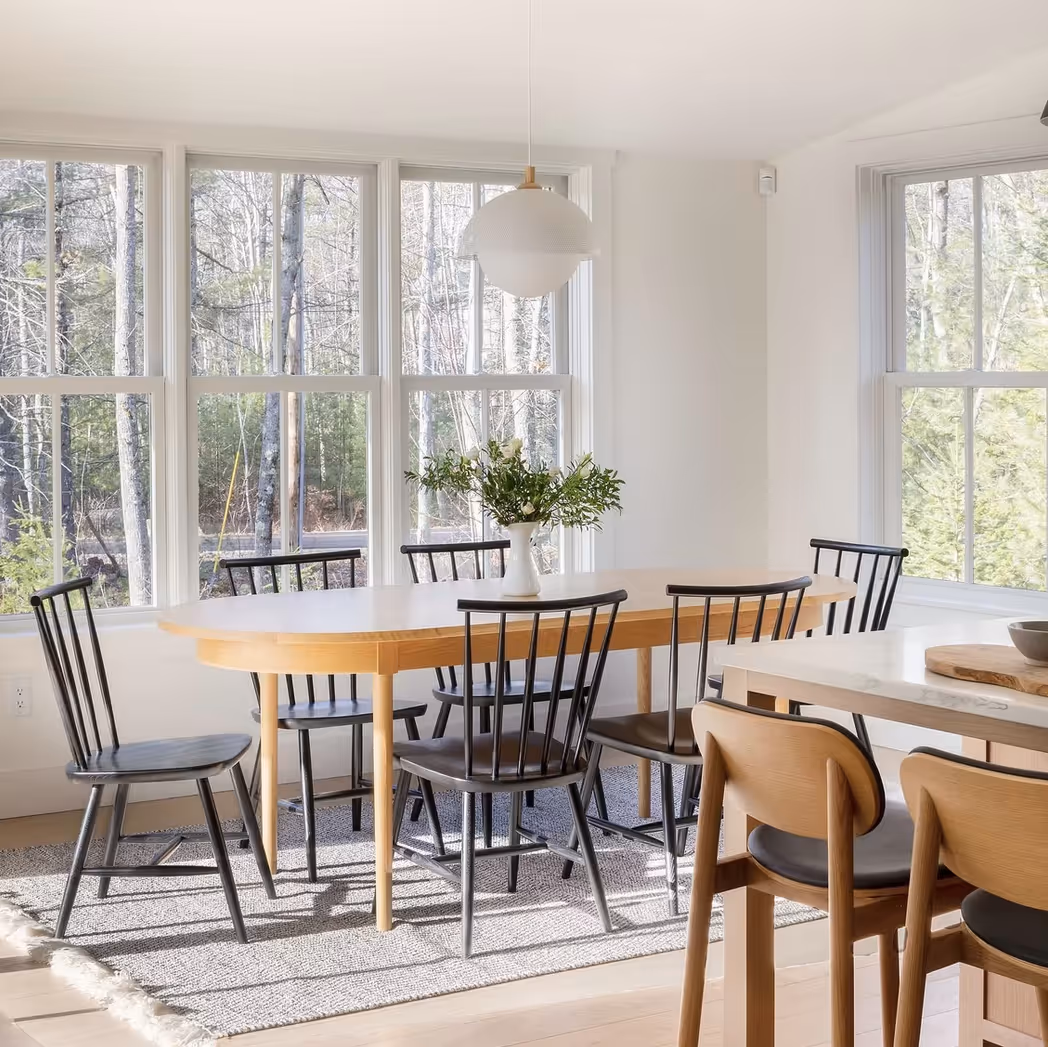 Bright dining area with a light wood oval table surrounded by six black chairs, a vase with greenery, large windows showing trees outside, and part of a kitchen island with two wooden stools.