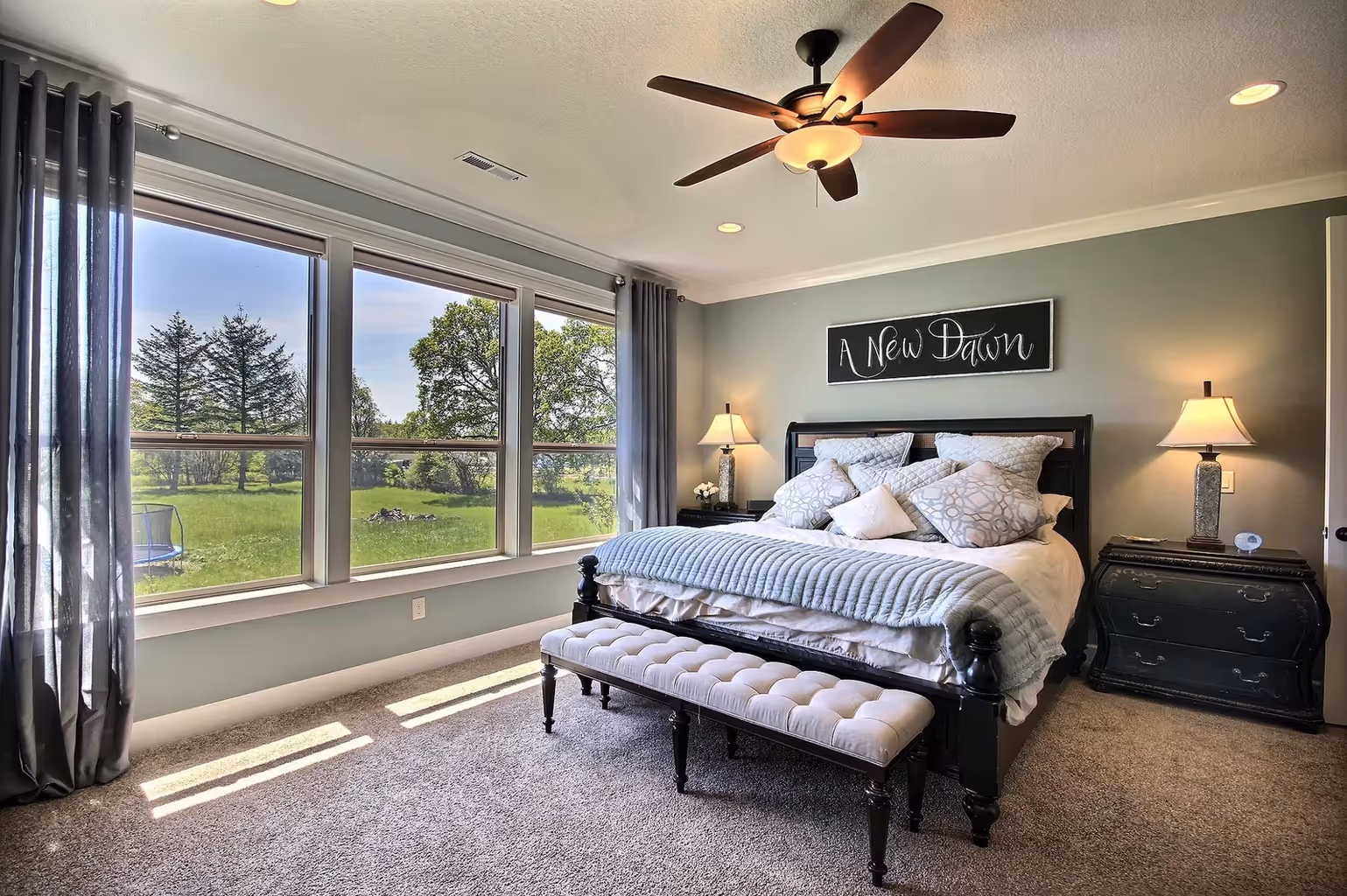 Bright bedroom with large windows, a black bed frame with multiple pillows and a bench, bedside tables with lamps, and a ceiling fan overhead.