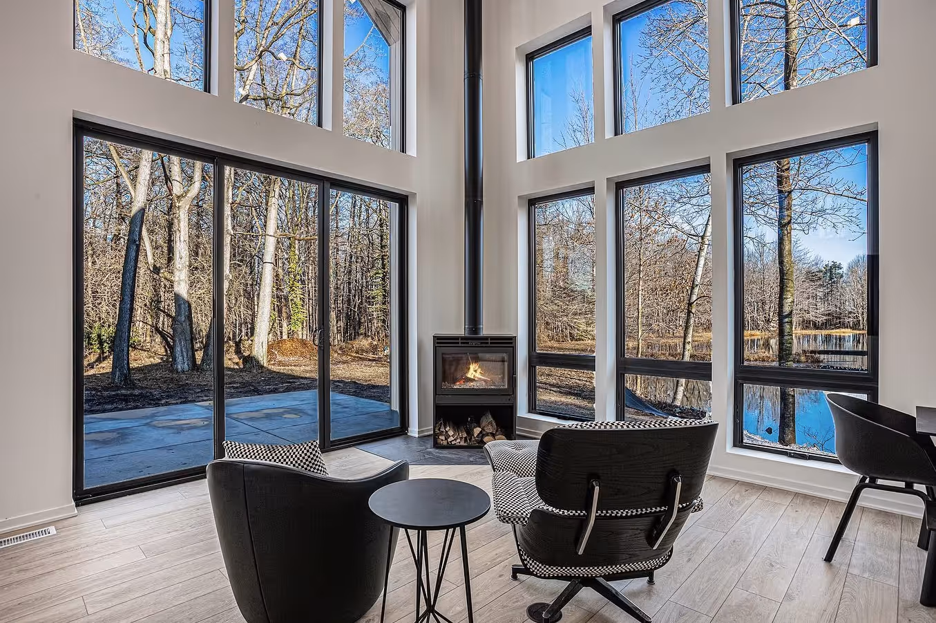 Modern living room with large floor-to-ceiling windows showing a wooded outdoor view, a fireplace, two black and white armchairs, and a small round table.