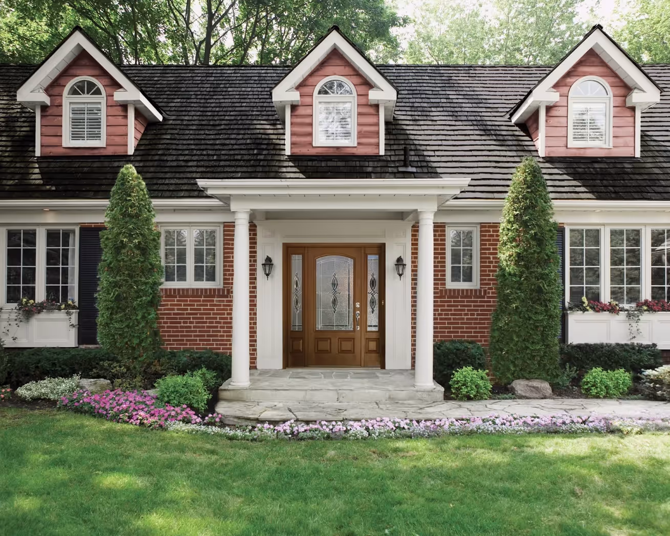 Front view of a brick house with three dormer windows, a wooden door with decorative glass, stone steps, and landscaped garden with tall shrubs and pink flowers.