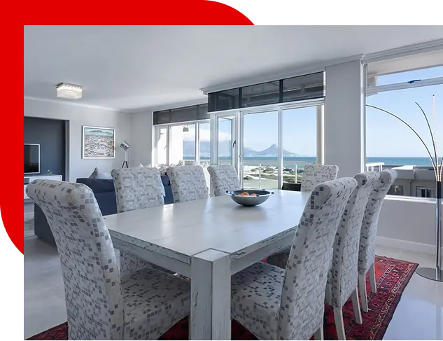 Modern dining room with a large white table surrounded by eight patterned chairs, a red rug underneath, large windows with a scenic mountain and ocean view, and a tall floor lamp in the corner.