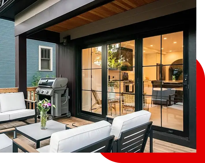 Outdoor patio with white cushioned chairs, a coffee table with flowers, and a stainless steel grill next to large glass doors opening into a modern kitchen.