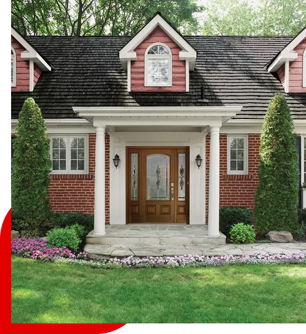 Red brick house entrance with wooden door, white columns, dormer windows, tall shrubs, and flowerbeds on a green lawn.