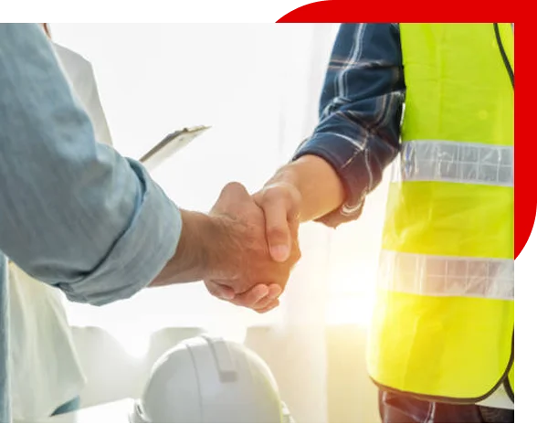 Two people shaking hands, one wearing a yellow safety vest and a plaid shirt, with a white safety helmet on a desk nearby.