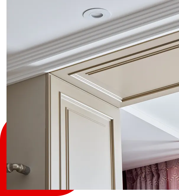 Corner view of beige kitchen cabinetry with decorative paneling and crown molding under a white ceiling with a recessed light.