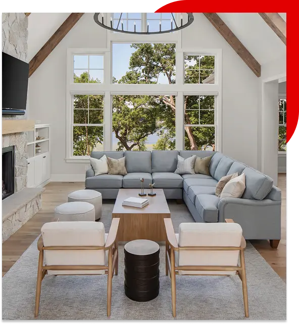Modern living room with light blue sectional sofa, wooden armchairs, and large windows showing green trees outside.