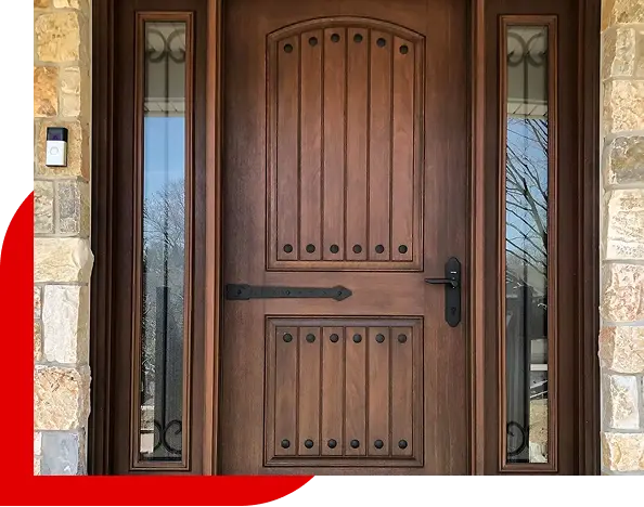 Solid wooden front door with vertical panel design and metal studs, flanked by two narrow glass windows with wrought iron accents, set in a stone wall.