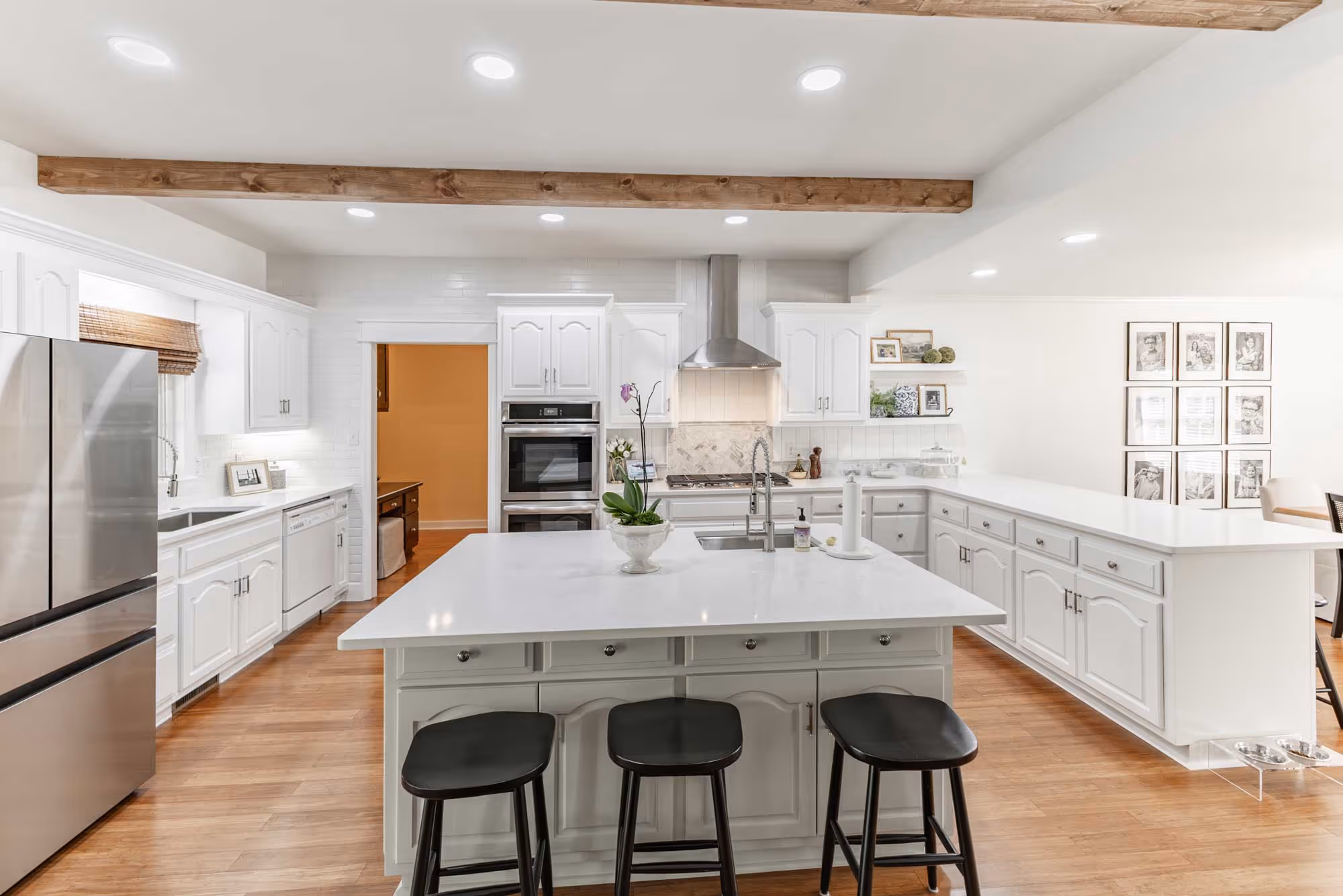 Bright modern kitchen with white cabinets, a large island with three black stools, stainless steel appliances, and wooden ceiling beams.