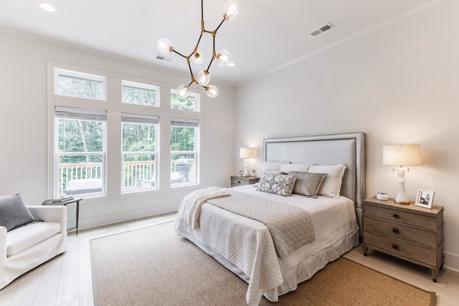Bright bedroom with large windows, a bed with gray and white bedding, two bedside tables with lamps, and a white armchair.