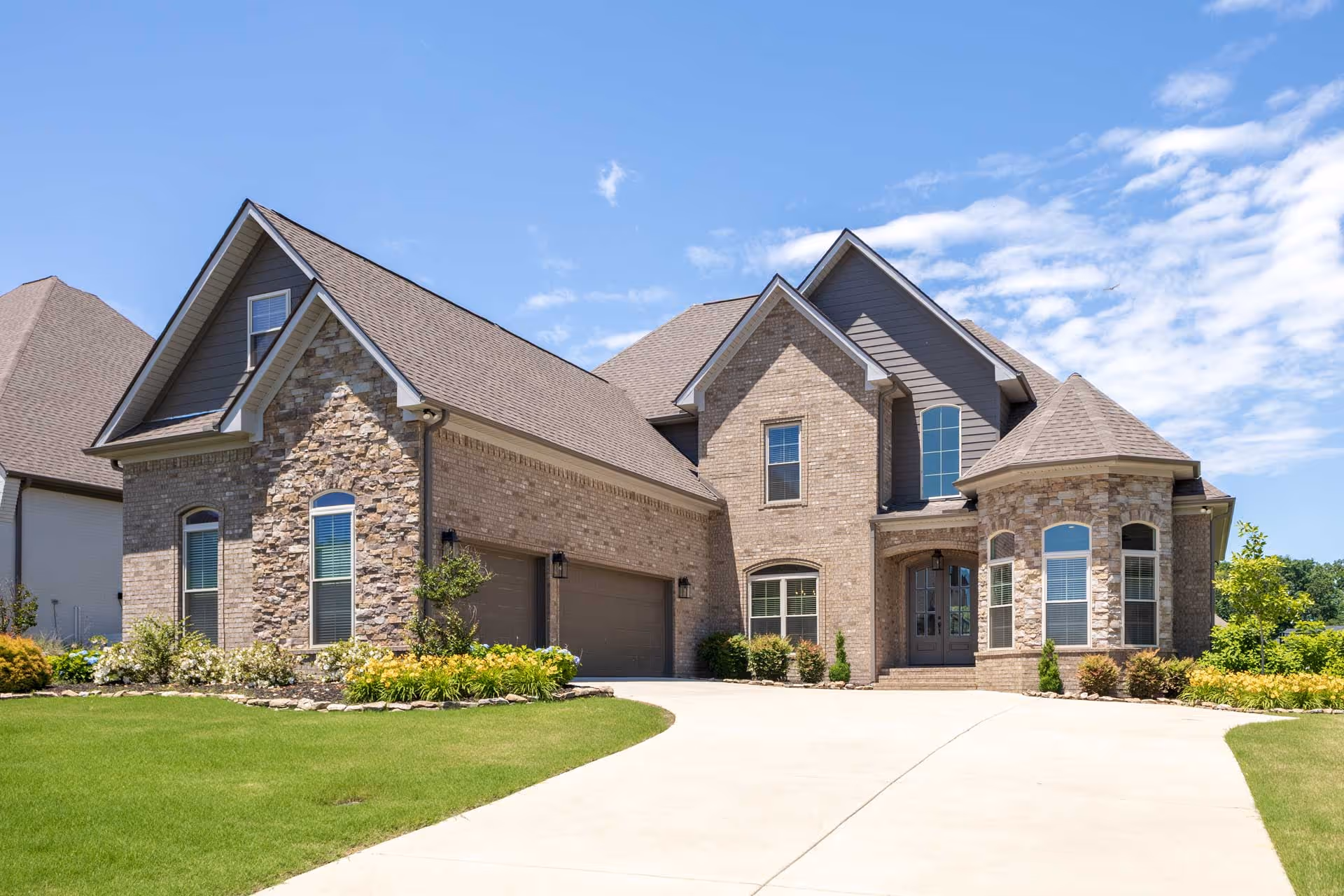 Large two-story brick and stone house with a three-car garage, arched windows, and landscaped lawn under a blue sky.