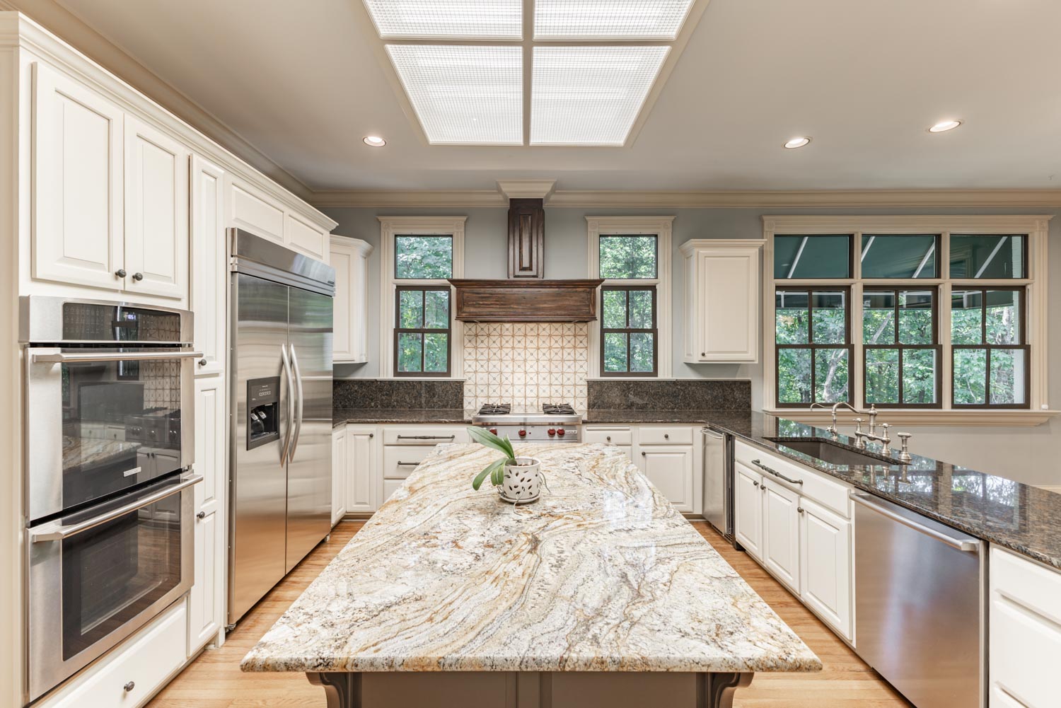Modern kitchen with marble island, stainless steel appliances, white cabinets, and large windows overlooking greenery.