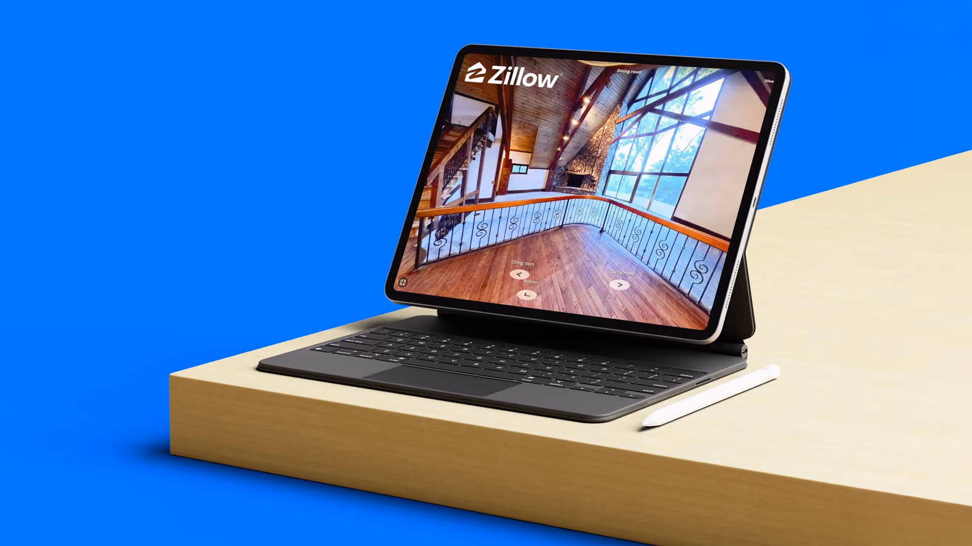Tablet with keyboard and stylus on wooden table showing a Zillow 3D home tour of a spacious room with wooden floors and large windows.
