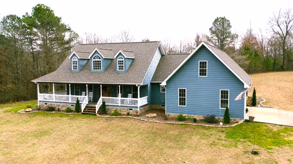 Blue two-story house with a large front porch and dormer windows, surrounded by grass and trees.