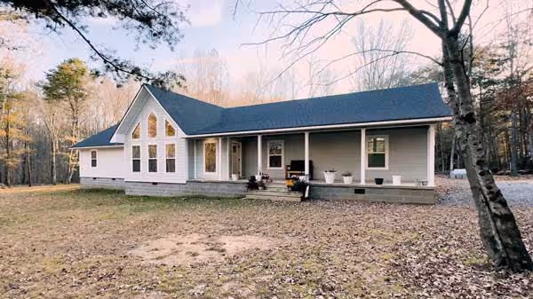 Single-story house with large triangular windows and a front porch, surrounded by trees in a woodland setting.