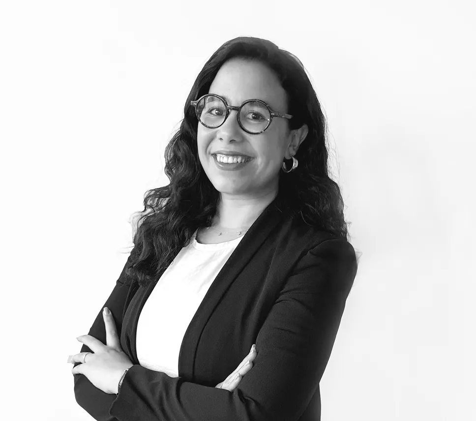 Portrait en noir et blanc d'une femme souriante avec des lunettes, les bras croisés, portant un blazer noir et un t-shirt blanc.