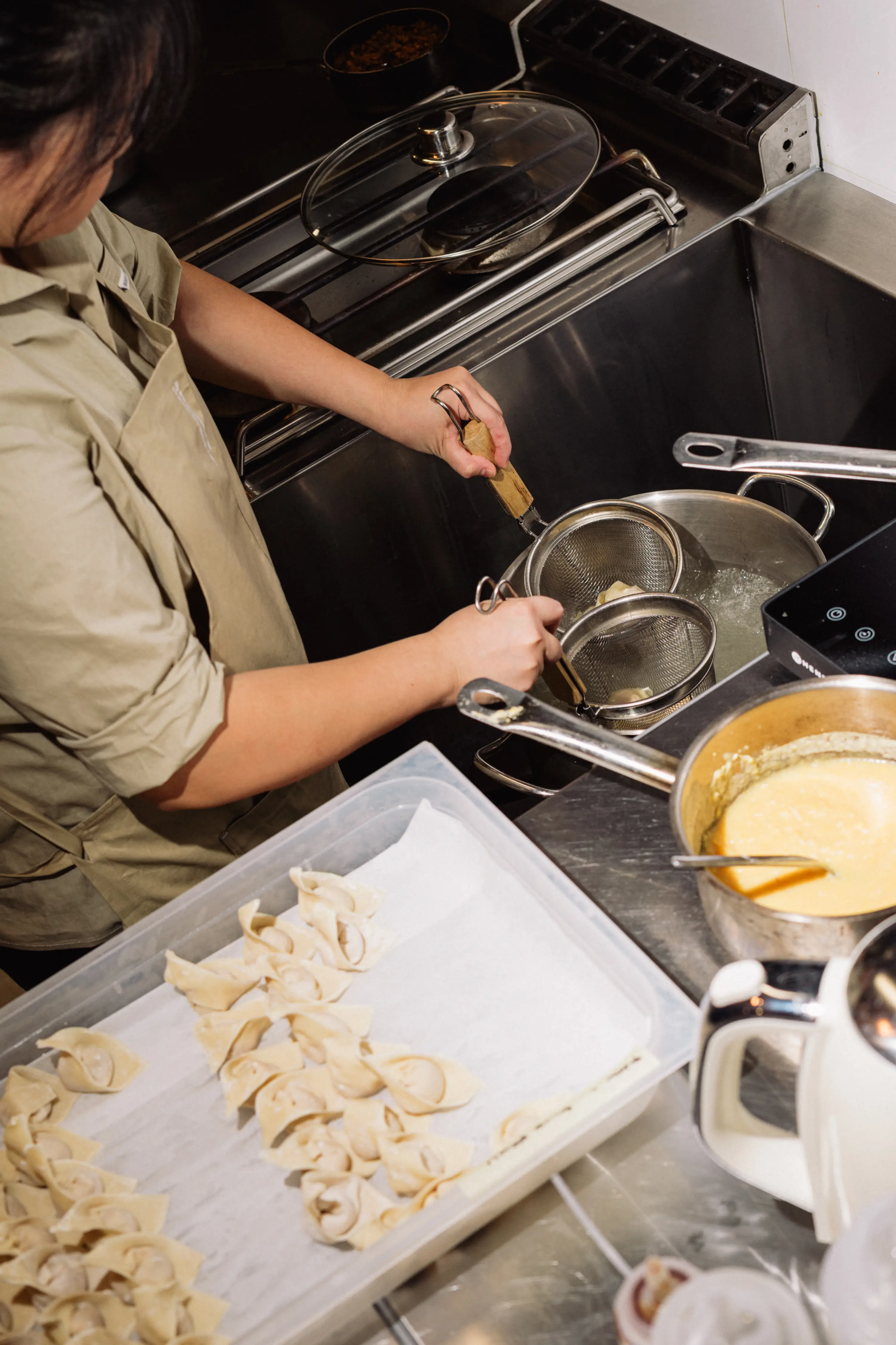 Person cooking dumplings using wire strainers over a pot of boiling water next to a tray of uncooked dumplings and a saucepan with sauce.