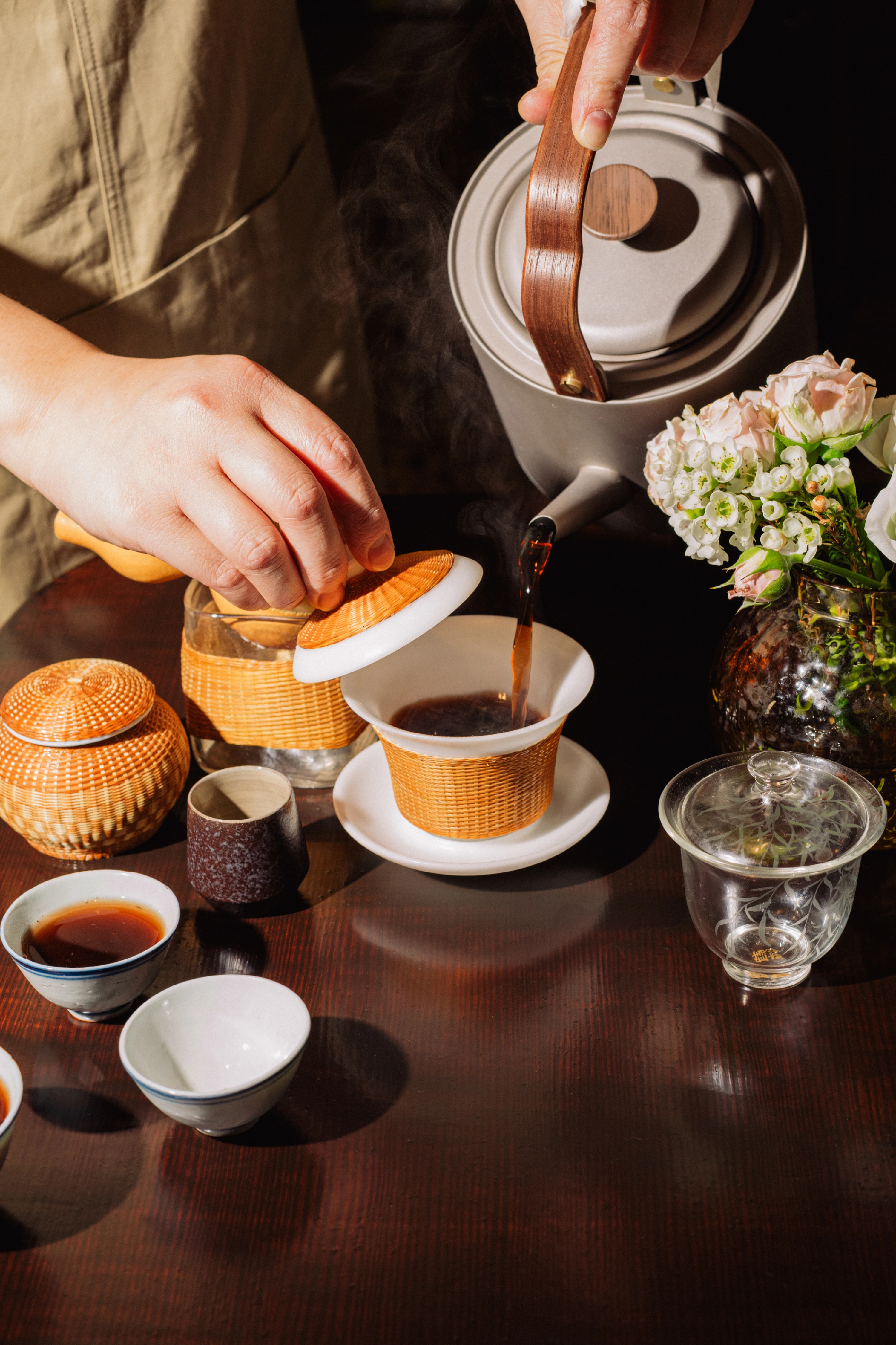 Person lifting lid of ceramic cup as dark tea is poured from a kettle into it on a wooden table with additional tea cups and a vase of flowers.