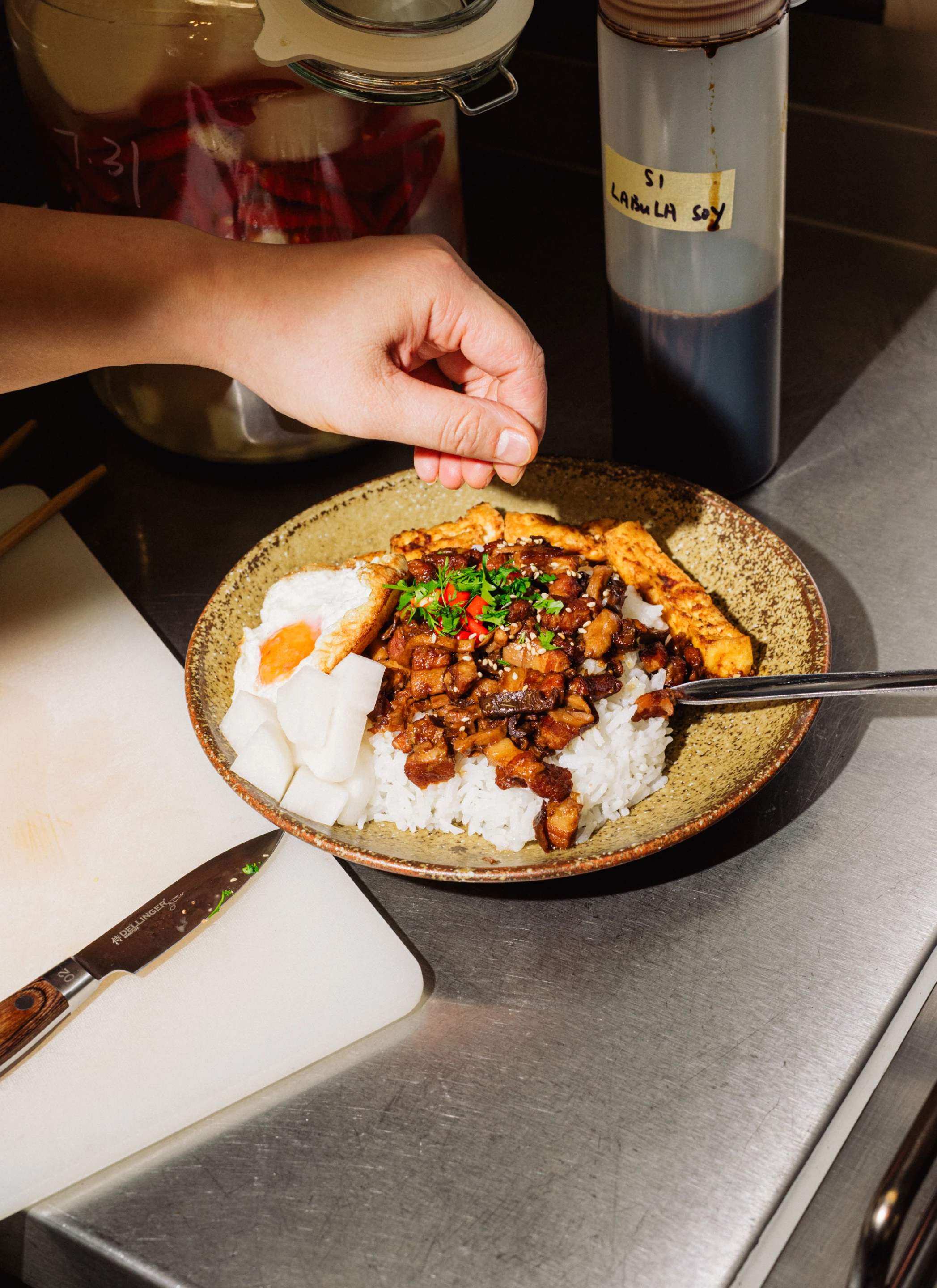 Hand sprinkling garnish over a bowl of rice topped with diced meat, a fried egg, tofu strips, and pickled radish on a stainless steel countertop.