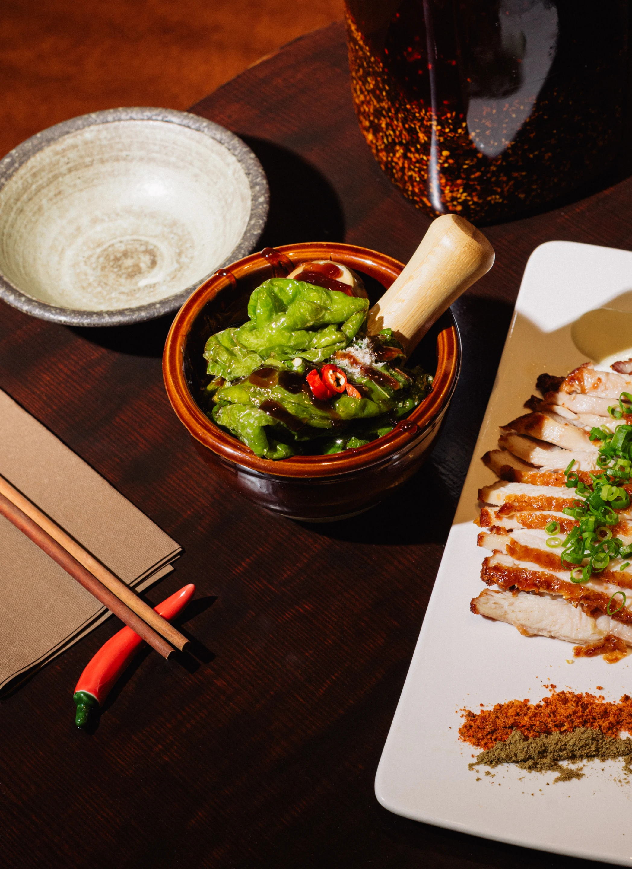 Sliced grilled chicken topped with chopped green onions and two spice powders on a white rectangular plate beside a ceramic bowl with lettuce and red chili garnished with sauce.