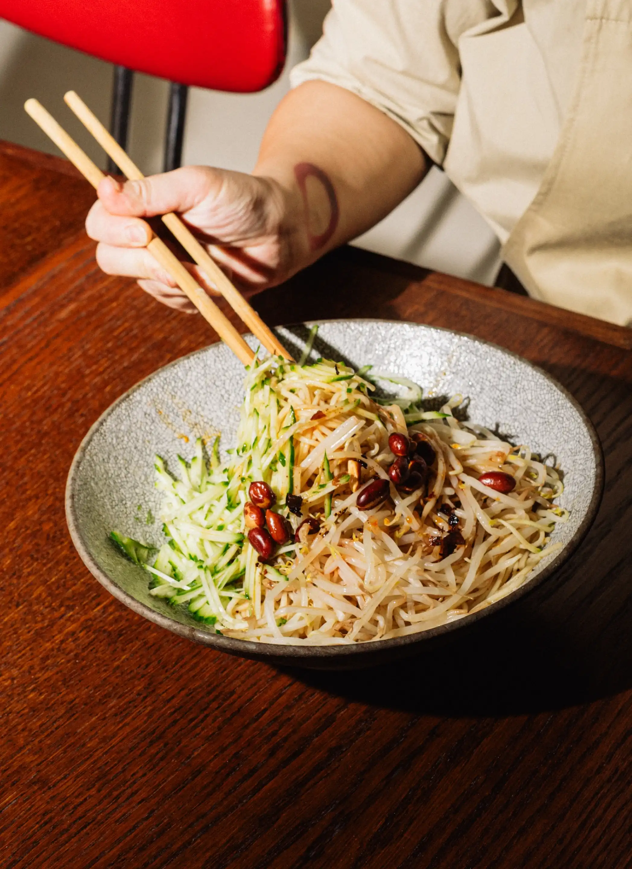 Person holding chopsticks picking up noodles mixed with bean sprouts, cucumber, and peanuts in a gray bowl on a wooden table.